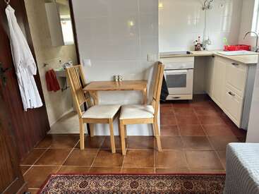 A cozy kitchen with white cabinets, a small wooden table with two chairs, brown tile floor, an adjacent bathroom, and a patterned rug in the foreground.