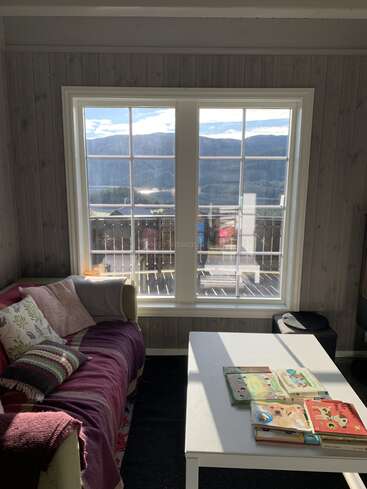 Sunlight streams through large windows, illuminating a cozy room with a purple-covered sofa, colorful pillows, white coffee table, and children's books. Scenic mountains are visible outside.