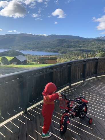 A young child in a red outfit stands on a wooden deck, holding a small bicycle. Behind, a scenic landscape features hills, lake, trees, and scattered houses.