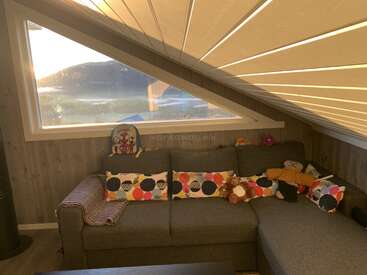 A cozy attic room with a slanted ceiling, a large window overlooking mountains, a grey sectional sofa, colorful cushions, and children’s toys scattered around.