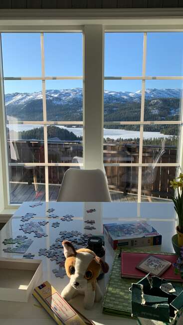 A sunlit room with a puzzle in progress, stuffed dog, and plants on a table. Through large windows, snowy mountains and a lake create a beautiful view.