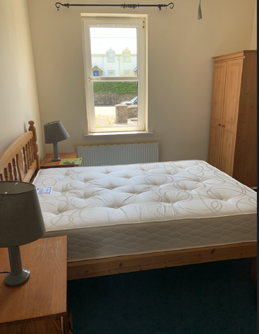 Une petite chambre avec un lit en bois, un matelas neuf, deux tables de chevet avec des lampes, une fenêtre, une armoire, un radiateur et une vue sur les maisons à l'extérieur.