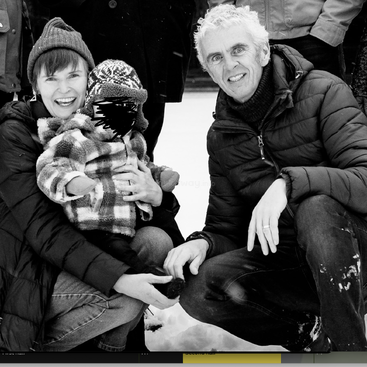 A imagem é uma fotografia em preto e branco de uma família de três pessoas posando para uma foto na neve, com o pai agachado e segurando o filho.
