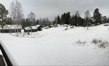 Eine friedliche Winterlandschaft im Dorf. Schnee bedeckt den Boden und die Dächer. Kahle Bäume und immergrüne Wälder umgeben rustikale Holzhäuser unter einem bewölkten, bedeckten Himmel.