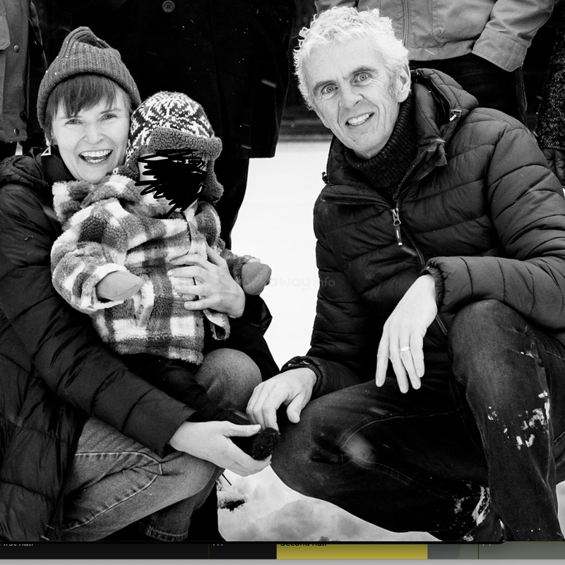 The image is a black-and-white photograph of a family of three posing for a photo in the snow, with the father crouching and holding his child.