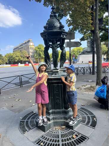 Two children pose cheerfully near an ornate green water fountain in a city square. Surrounding them are trees, a grand building, and a monument in the background.