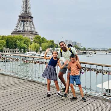 Un hombre y dos niños posan felices en un puente de París, con la Torre Eiffel y el río de fondo, rodeados de coloridos candados del amor.