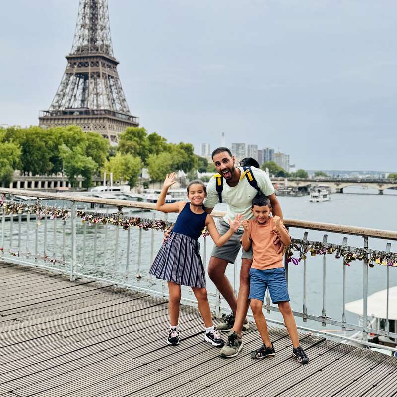 A man and two children pose happily on a bridge in Paris, with the Eiffel Tower and river in the background, surrounded by colorful love locks.