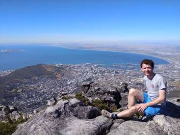 The image depicts a man sitting on a rocky outcrop overlooking a city and body of water, set against a clear blue sky.