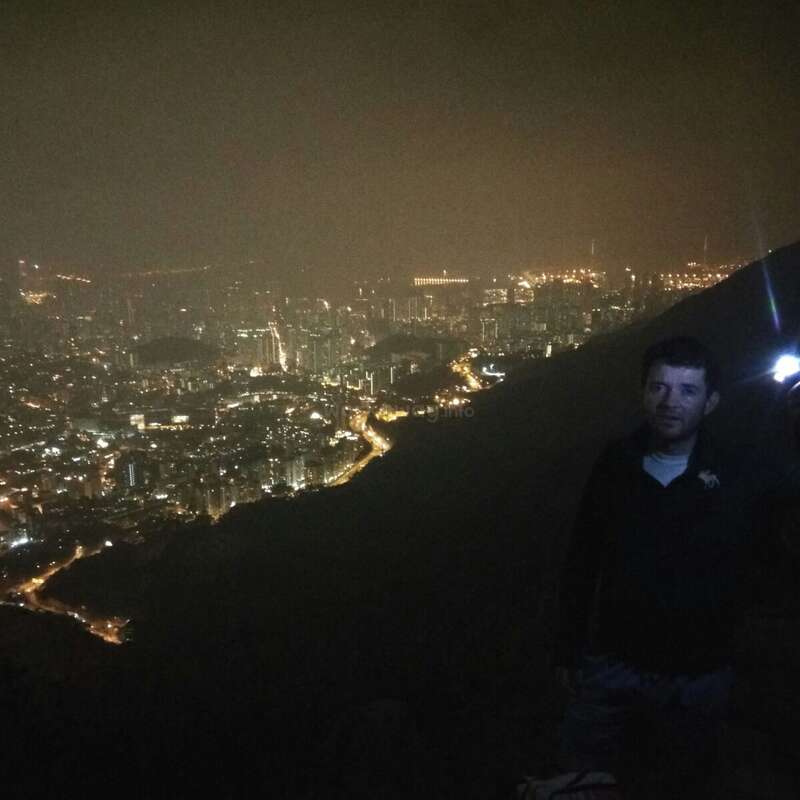 The image depicts a man standing on a mountain at night, with a cityscape below him, illuminated by streetlights and buildings, set against a dark sky.