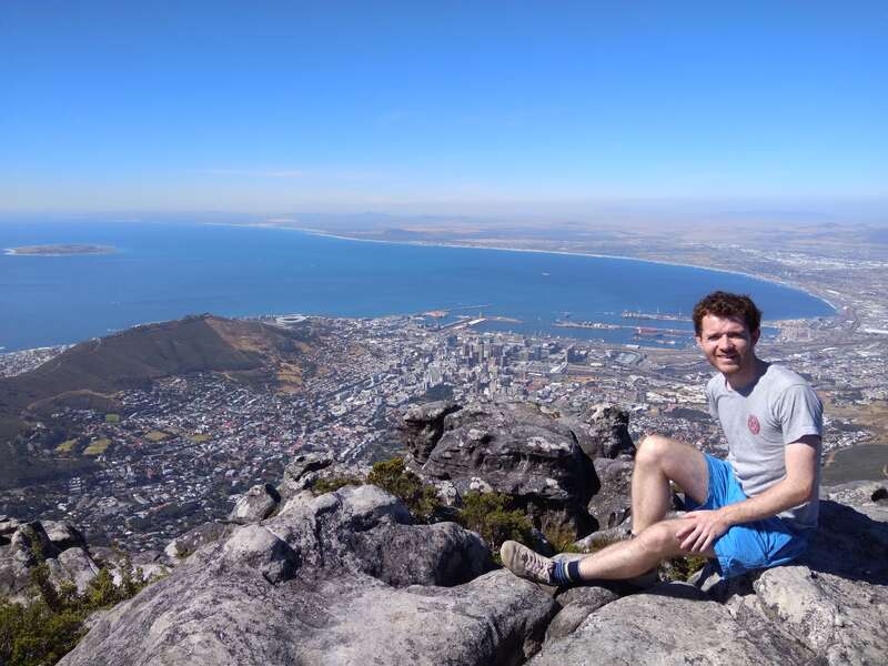 The image depicts a man sitting on a rocky outcrop overlooking a city and body of water, set against a clear blue sky.