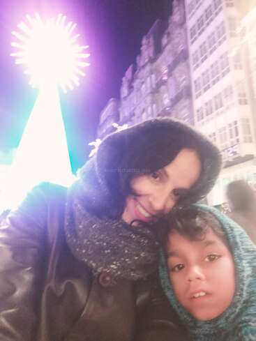 A woman and child bundled in winter clothes smile for a selfie at night, with a brightly lit Christmas tree and tall buildings in the background.