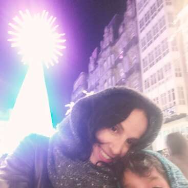 Eine Frau und ein Kind in Winterkleidung lächeln für ein nächtliches Selfie, während im Hintergrund ein hell erleuchteter Weihnachtsbaum und hohe Gebäude zu sehen sind.