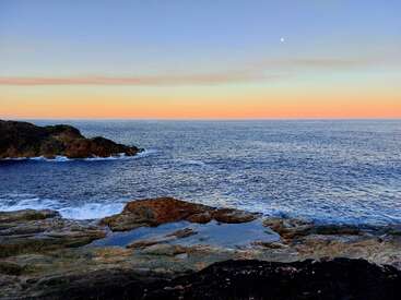 Un tranquilo paisaje marino al atardecer con suaves olas, rocas escarpadas y un cielo tranquilo iluminado por la luna con tonos naranjas y azules, creando una vista costera pacífica y pintoresca.