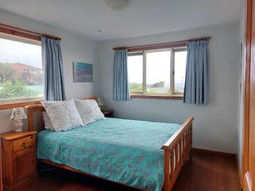 Esta acogedora habitación cuenta con una cama de madera, ropa de cama turquesa, almohadas blancas, mesitas de noche a juego, cortinas azul claro y grandes ventanas que proporcionan abundante luz natural.