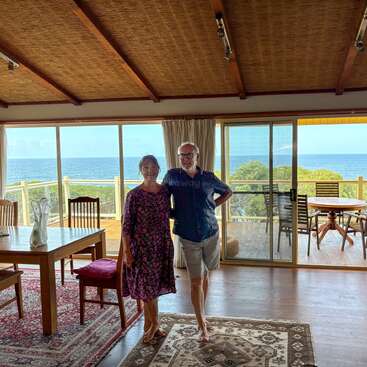 Ein lächelndes Paar steht in einem gemütlichen, sonnigen Zimmer mit Holzdecken und Teppichen und hat durch große Glasfenster und Türen einen tollen Blick aufs Meer.