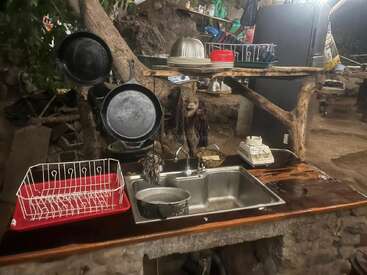This rustic outdoor kitchen features a metal sink, hanging cast iron pans, a dish rack, old telephone, wooden shelves, various dishes, and natural tree elements throughout.
