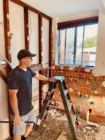 A man stands in a room undergoing renovation, holding a tool and leaning on a ladder, surrounded by exposed brick and debris.