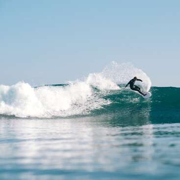 A person in a wetsuit is surfing on a wave with a white surfboard, creating a large splash of water. The sky is blue and clear.