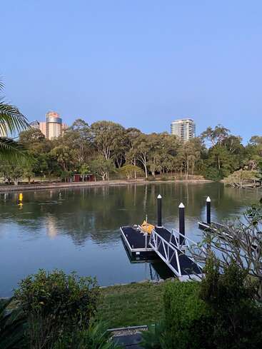 The image depicts a serene lake scene with a dock and a city skyline in the background, surrounded by lush trees and bushes on a clear day.