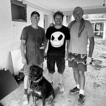 The image depicts three men standing together with a dog, posing for a photo in a garage or workshop setting, with a house visible in the background.
