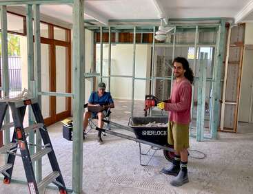 The image depicts two men in a room under construction, with one standing beside a wheelbarrow and the other seated on a chair, surrounded by building materials.