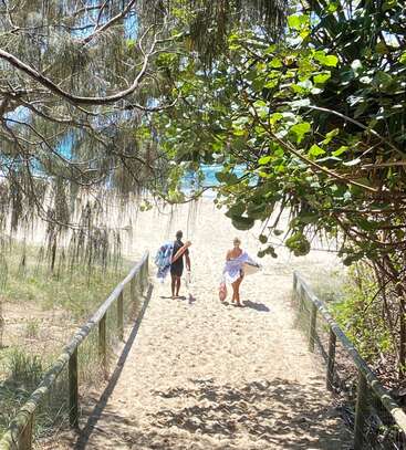 The image depicts a sandy path leading to the beach, flanked by a fence and trees, with two people walking towards the ocean, carrying surfboards.