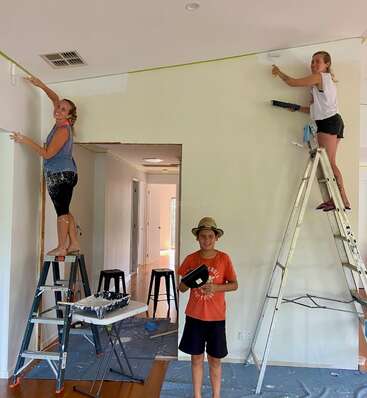 The image shows three people painting a room, with two on ladders and one standing in front, holding a tablet, amidst scattered paint supplies and a ladder.