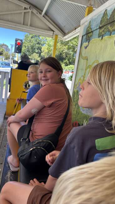 Three children sit on a bench at a covered outdoor area, smiling and waiting. There is colorful wall art, greenery, and a traffic light visible outside.