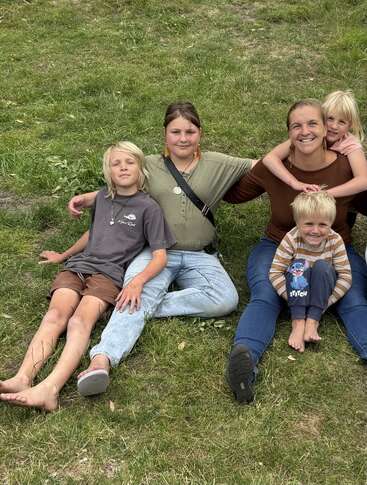 A woman and four children sit on green grass, smiling and relaxing together. They seem happy and close, enjoying a casual, outdoor family moment in nature.