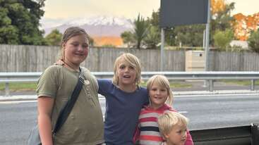 Four smiling children stand together on a roadside, with trees, a wooden fence, and a snow-capped mountain in the background under a pastel sky.