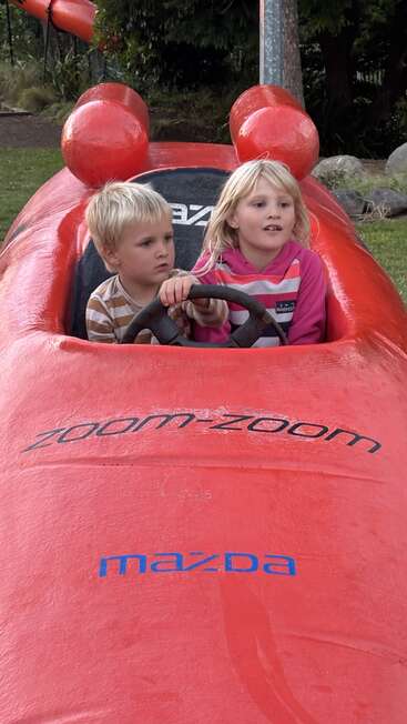 Two young children sit together in a bright red Mazda-themed play car outdoors, looking ahead with interest. "Zoom-Zoom" is written on the front. Playful scene.