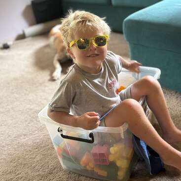 A smiling young boy wearing yellow sunglasses sits inside a plastic container filled with colorful toy blocks on a carpeted floor. A dog lies in the background.