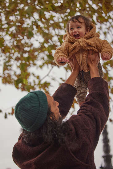 Das Bild zeigt einen Mann in einem braunen Pullover und einer grünen Mütze, der ein Baby in einem hellbraunen Mantel in die Luft hebt, mit einem Baum im Hintergrund.