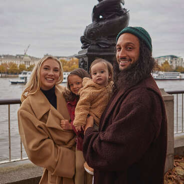 Das Bild zeigt eine vierköpfige Familie, die an einem Fluss steht. Die Mutter und der Vater halten ihre beiden kleinen Kinder im Arm, alle sind warm angezogen und lächeln in die Kamera.