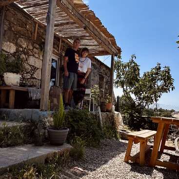 Ein Mann und eine Frau stehen lächelnd auf einer rustikalen Steinveranda, umgeben von Topfpflanzen, mit einem hölzernen Picknicktisch und einem sonnigen blauen Himmel im Hintergrund.