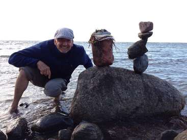 L'image représente un homme accroupi à côté d'une grande formation rocheuse sur une plage, avec une pile de pierres en équilibre sur le dessus, sur fond d'océan serein.