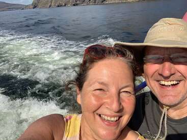 L'image montre un homme et une femme posant pour un selfie sur un bateau, avec l'océan et la côte rocheuse visibles à l'arrière-plan.