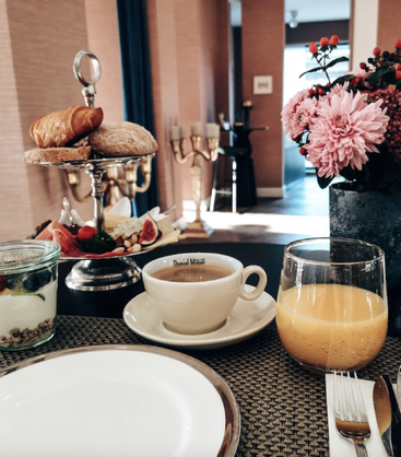 Une table de petit-déjeuner joliment dressée avec des viennoiseries, du café, du jus d'orange, un parfait au yaourt, des fleurs fraîches et des assiettes élégantes dans une salle à manger accueillante et chaleureusement éclairée.
