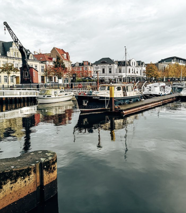 Scène de port paisible avec divers bateaux amarrés, des bâtiments historiques colorés, des arbres d'automne, une grue et leurs reflets qui scintillent doucement sur l'eau calme. Ciel nuageux.