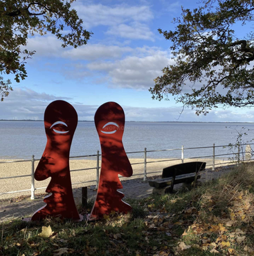 Deux sculptures abstraites rouges en forme de visage se trouvent au bord d'une rivière paisible, près d'un banc. Des feuilles d'automne jonchent le sol, tandis que des arbres encadrent un ciel bleu et l'eau.