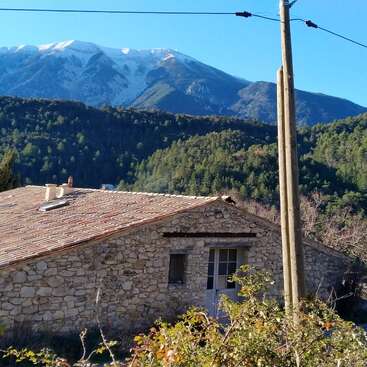 Uma casa rústica de pedra fica aninhada entre a vegetação e os arbustos. Atrás dela, colinas arborizadas se erguem em camadas, com montanhas cobertas de neve se elevando majestosamente sob um céu azul límpido.