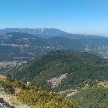 Uma paisagem montanhosa deslumbrante sob um céu azul límpido, com colinas verdes em camadas, florestas densas, picos distantes e campos espalhados lá embaixo. Uma vista tranquila.