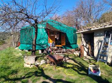 Uma estrutura verde parecida com uma barraca fica ao lado de um galpão de pedra com uma escada, árvores e ferramentas de jardim. A luz do sol ilumina a cena, criando uma atmosfera rústica e campestre.