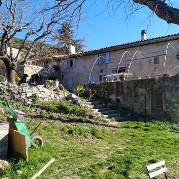 Essa imagem mostra uma casa rústica de pedra com venezianas azuis, um jardim em terraços, árvores sem folhas, grama verde, ferramentas de jardinagem espalhadas e um céu azul claro e ensolarado.