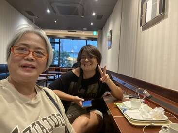Two women are sitting in a cozy cafe. One smiles at the camera, the other makes a peace sign. Used cups and plates are on the table.