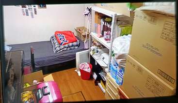 This image shows a small, tidy bedroom with a single bed, shelves, storage boxes, a red trash bin, pink suitcase, and personal belongings neatly organized.
