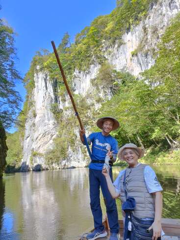 Two smiling people in hats enjoy a boat ride on a calm river, surrounded by steep cliffs, green trees, and a bright blue sky. Peaceful atmosphere.