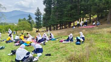 This image shows many students, mostly girls in school uniforms, studying and relaxing outdoors on the grass, surrounded by trees and mountains on a clear day.