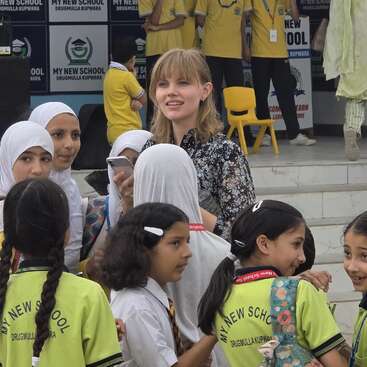 Eine Gruppe lächelnder Kinder, von denen einige Kopftücher und Schuluniformen tragen, umringt eine Frau mit einem Telefon in der "My New School" in Drugmulla Kupwara.